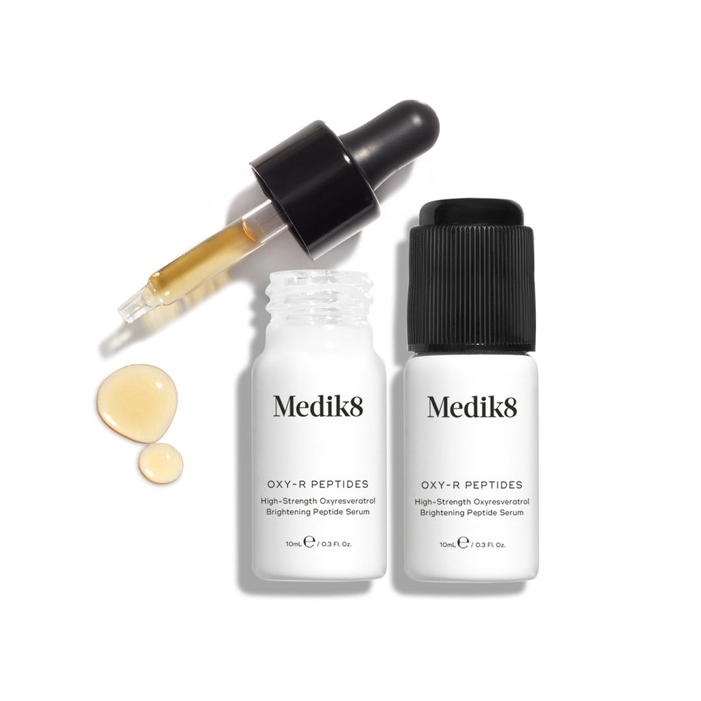 Dwie małe otwarte białe butelki serum Oxy-R Peptides są zaprezentowane na jasnym tle. Obok leży czarna pipeta, a na białej powierzchni widoczne są dwie krople żółtego serum.
