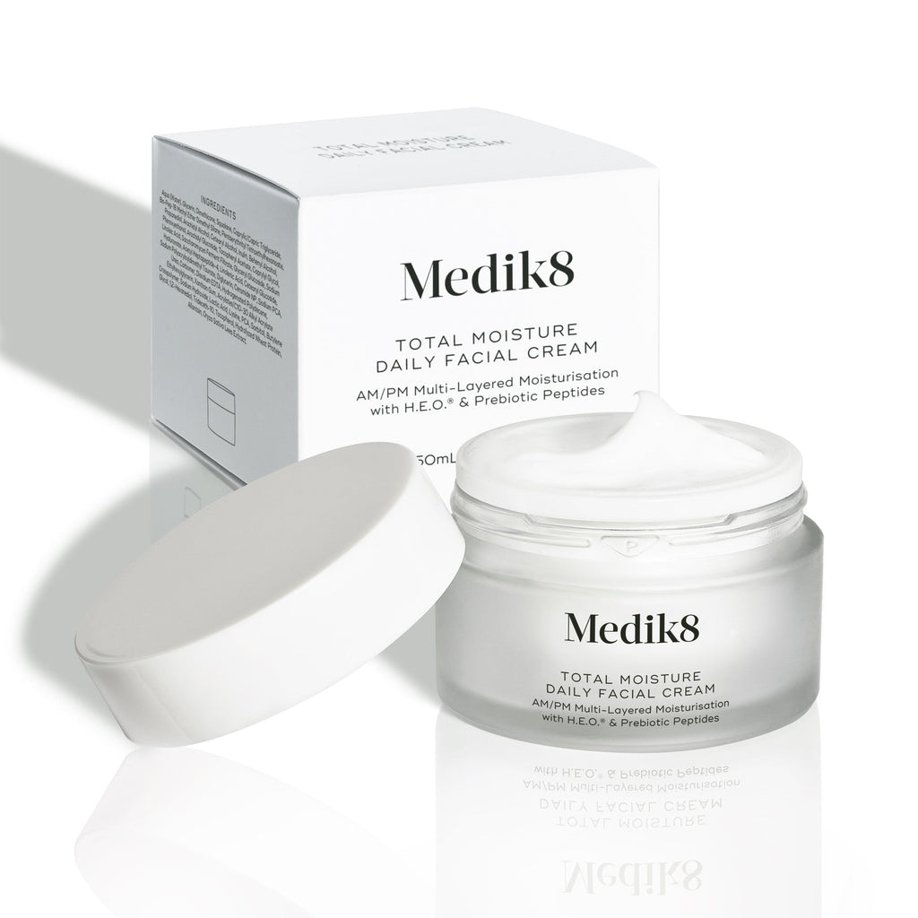 Otwarty matowy szklany słoiczek Medik8 Total Moisture Daily Facial Cream z białą zakrętką umieszczoną obok, prezentujący bogaty biały krem. Słoiczek stoi przed białym kartonowym opakowaniem na jasnym tle. Tekst na obu opakowaniach brzmi: „AM/PM wielowarstwowe nawilżenie z H.E.O.® i prebiotycznymi peptydami”.
