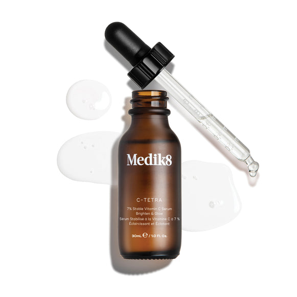 Ujęcie z góry typu packshot przedstawiające 30 ml butelkę Medik8 C-Tetra Lipid Vitamin C Radiance Serum. Bursztynowa szklana butelka jest otwarta i ustawiona centralnie, a czarna pipeta leży ukośnie za nią. Przezroczyste serum jest artystycznie rozlane i tworzy kałuże na czystym białym tle wokół butelki, z widocznymi kroplami na szklanej pipetcie. Oświetlenie jest jasne, rzuca miękkie cienie w lewo i tworzy kliniczny, a jednocześnie elegancki efekt wizualny.-14