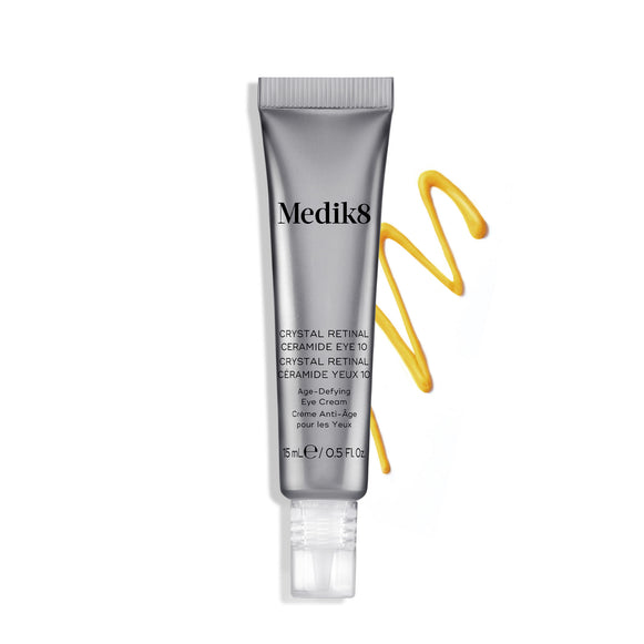 A silver tube of Crystal Retinal Ceramide Eye 10 stands upright, with a swirl of yellow-orange cream to its right.-hover-7