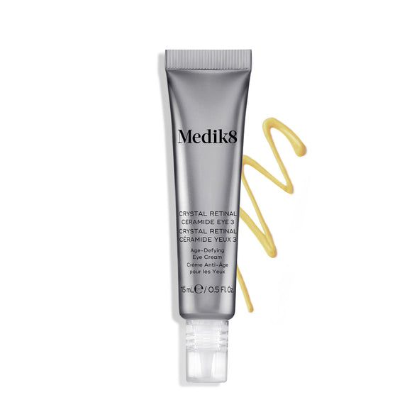 A silver tube of Crystal Retinal Ceramide Eye 3 stands upright, with a swirl of yellow-orange cream to its right.-7
