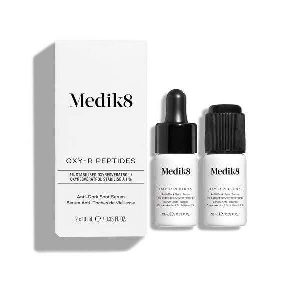 Dwie małe białe butelki serum Oxy-R Peptides są zaprezentowane na białym tle. Obok leży czarna pipeta, a na powierzchni widoczne są dwie krople żółtego serum.-33